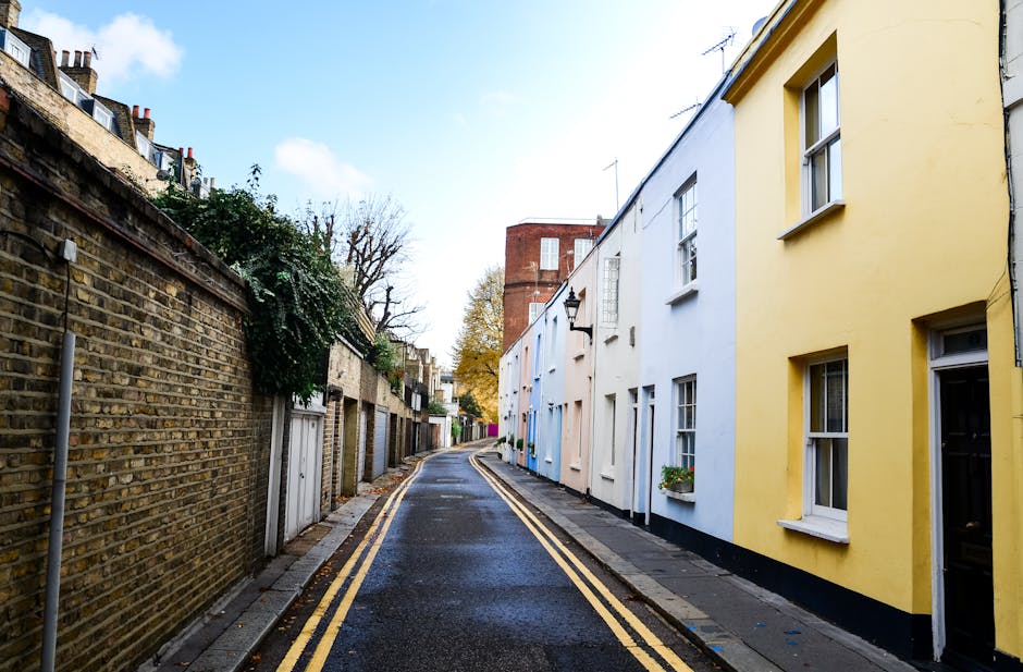 A black-and-white image of a workspace featuring an open laptop displaying lines of code on the screen, positioned on a dark wooden desk. To the right of the laptop, there is a white notebook with a b
