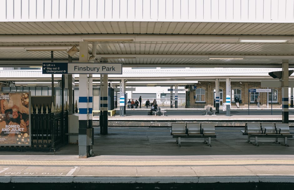 An outdoor tram stop with a covered waiting area featuring transparent roofing panels supported by metal posts. The platform has a metal railing and a digital timetable display, with a sign showing sa