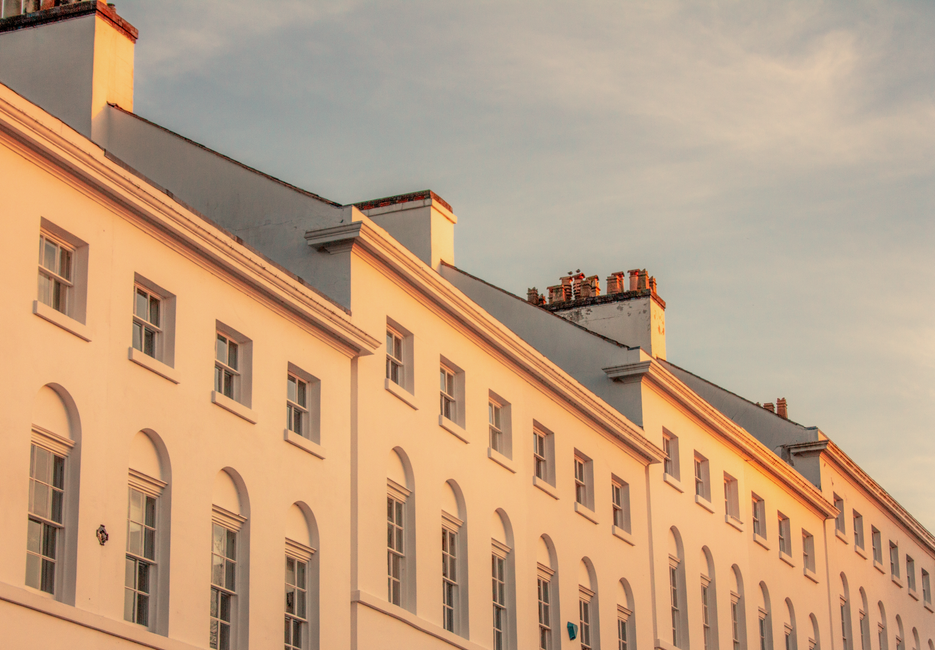 The image depicts a row of residential buildings with white facades and multiple rectangular windows, illuminated by warm sunlight during sunset or sunrise. The rooftops feature chimney stacks made of